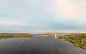 Zwischen Wollen und Dranbleiben Hängebrücke über einen breiten Fluss, umgeben von bewaldeten Ufern im warmen Abendlicht unter weitem Himmel.