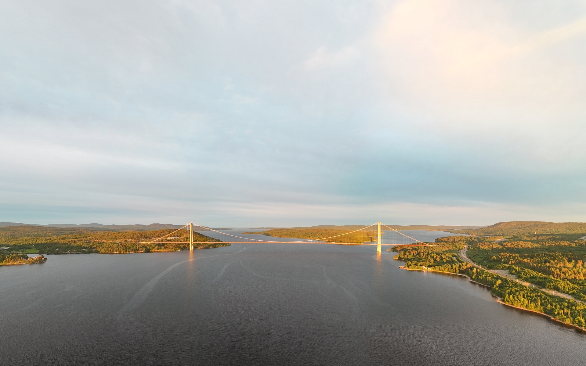 Hängebrücke über einen breiten Fluss, umgeben von bewaldeten Ufern im warmen Abendlicht unter weitem Himmel.